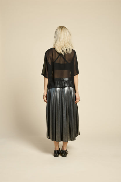Back view of  woman wearing the Feyer Skirt by Cokluch in Glow Knit Silver, a midi length pleated skirt in shimmery metallic fabric. She is wearing it with a sheer black graphic top and standing in front of a beige background. 