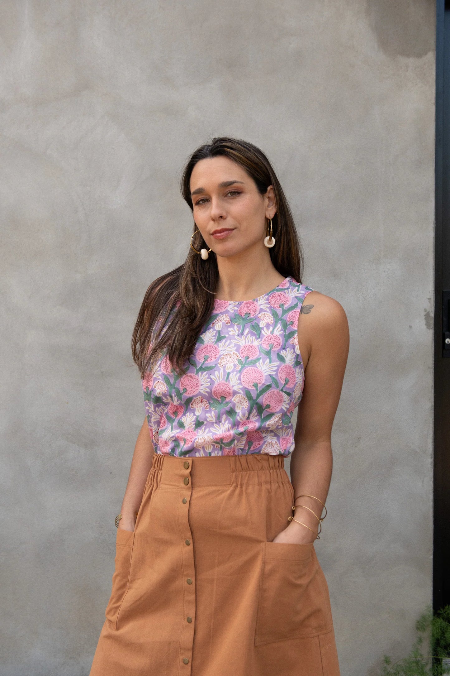 A woman wearing Forsythia top from Kazak in Lilac Floral, a cropped tank with a cutout and tie detail at the back. She is wearing it with the Nymphea Skirt in Ginger and standing in front of a grey wall. 