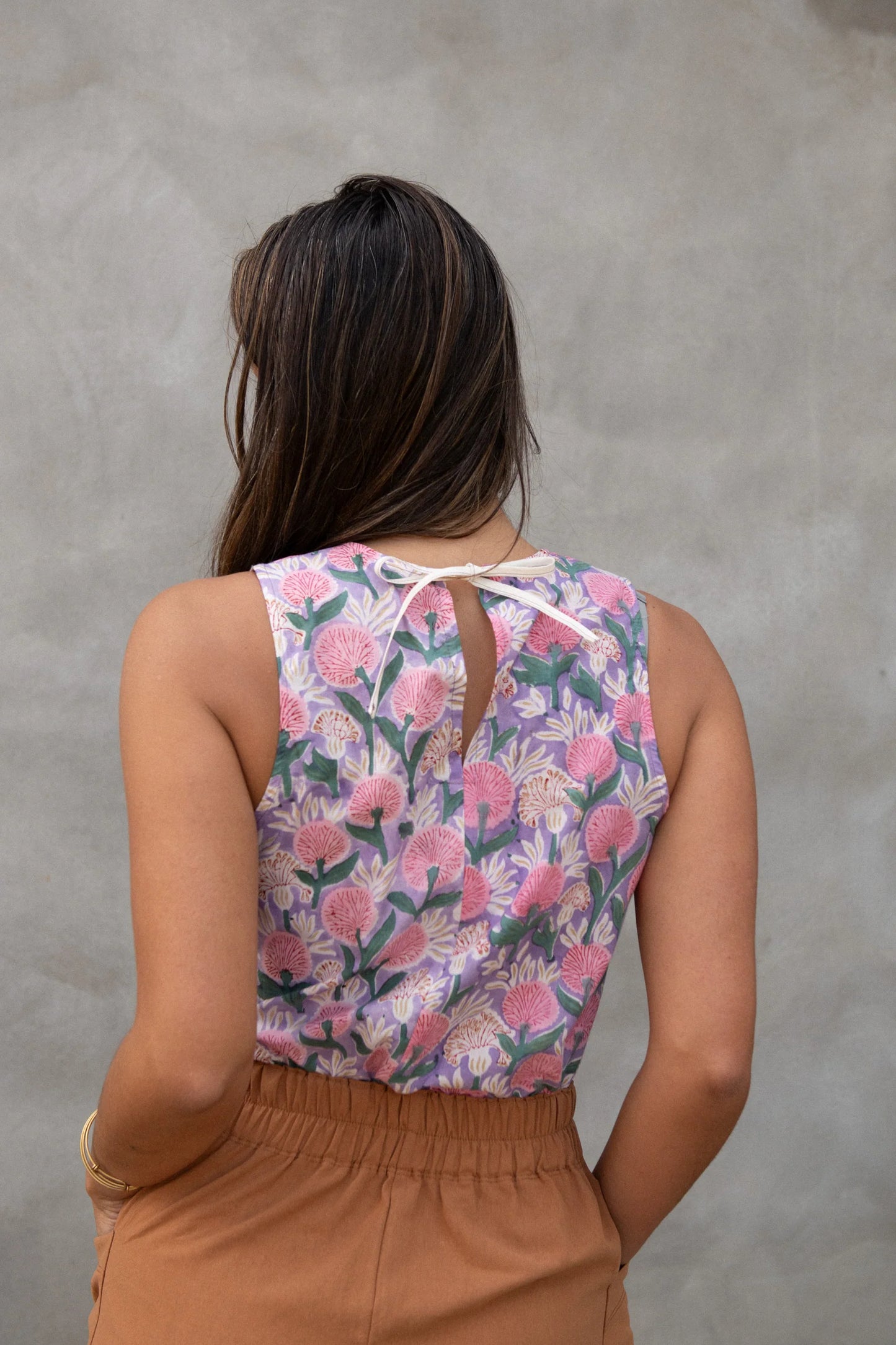 Back close up view of a woman wearing Forsythia top from Kazak in Lilac Floral, a cropped tank with a cutout and tie detail at the back. She is wearing it with the Nymphea Skirt in Ginger and standing in front of a grey wall. 