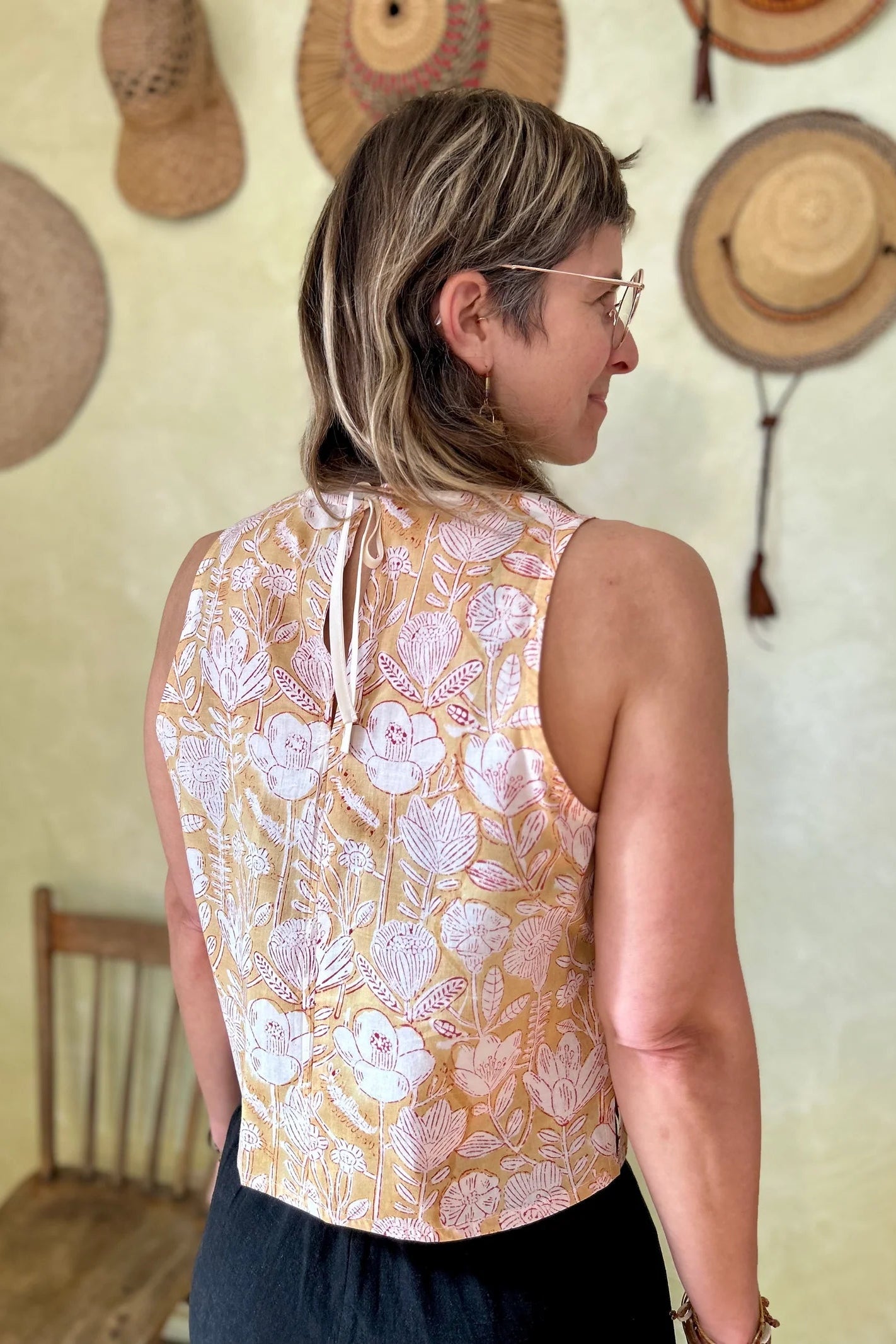Back view of a woman wearing Forsythia top from Kazak in Mustard Floral, a cropped tank with a cutout and tie detail at the back. She is wearing it with black ants and standing in front of a wall with hat hanging on it. 