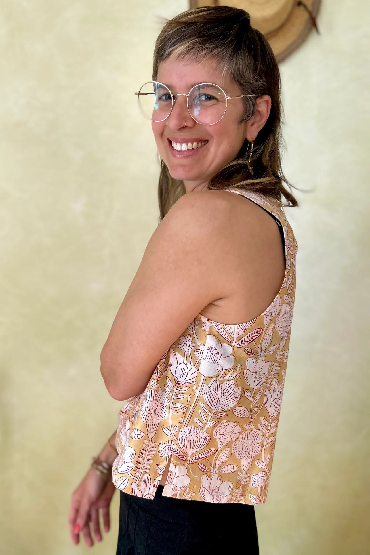 Side view of a woman wearing Forsythia top from Kazak in Mustard Floral, a cropped tank with a cutout and tie detail at the back. She is wearing it black pants and standing in of a chair and a wall with hats hanging on it. 