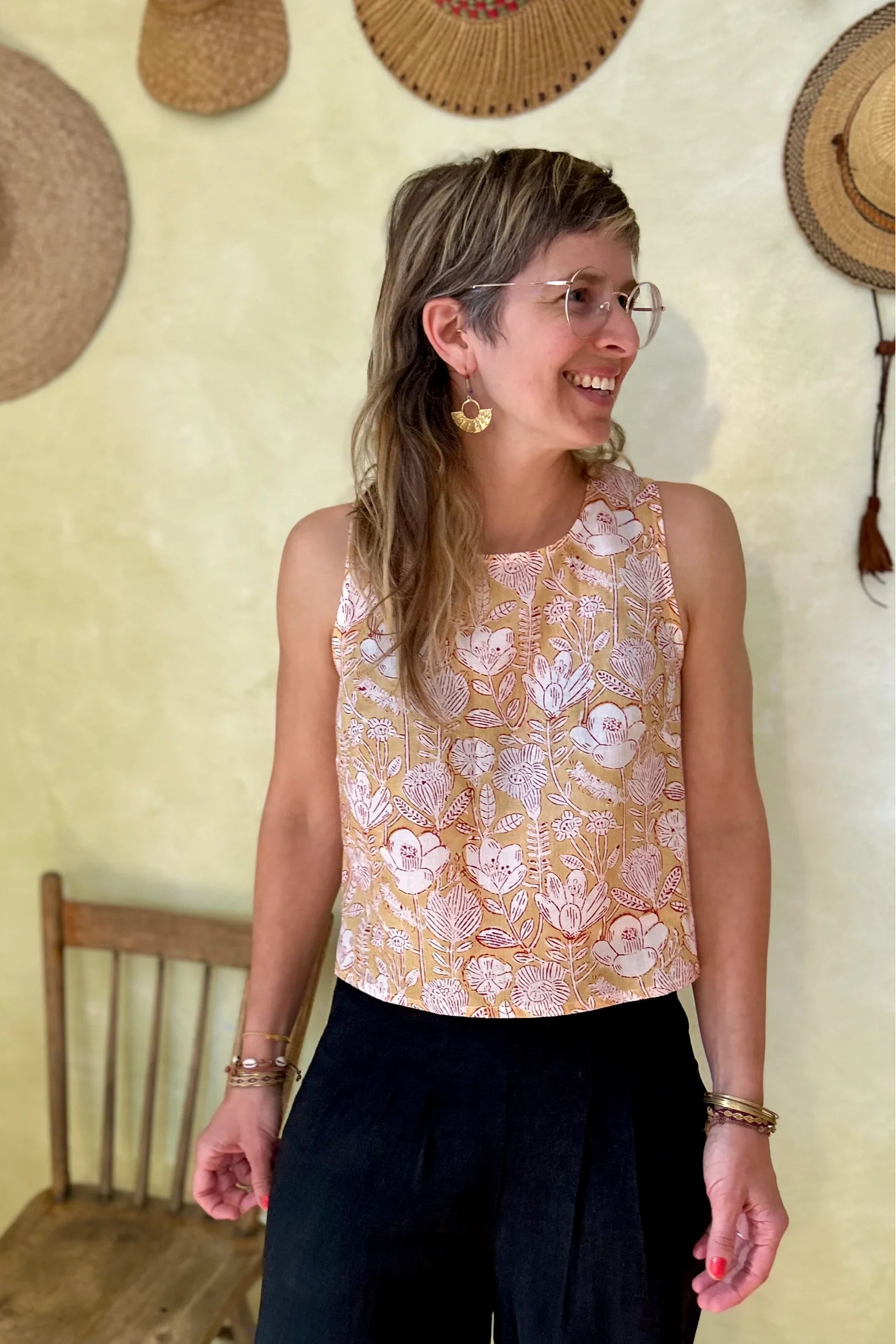 A woman wearing Forsythia top from Kazak in Mustard Floral, a cropped tank with a cutout and tie detail at the back. She is wearing it black pants and standing in of a chair and a wall with hats hanging on it. 