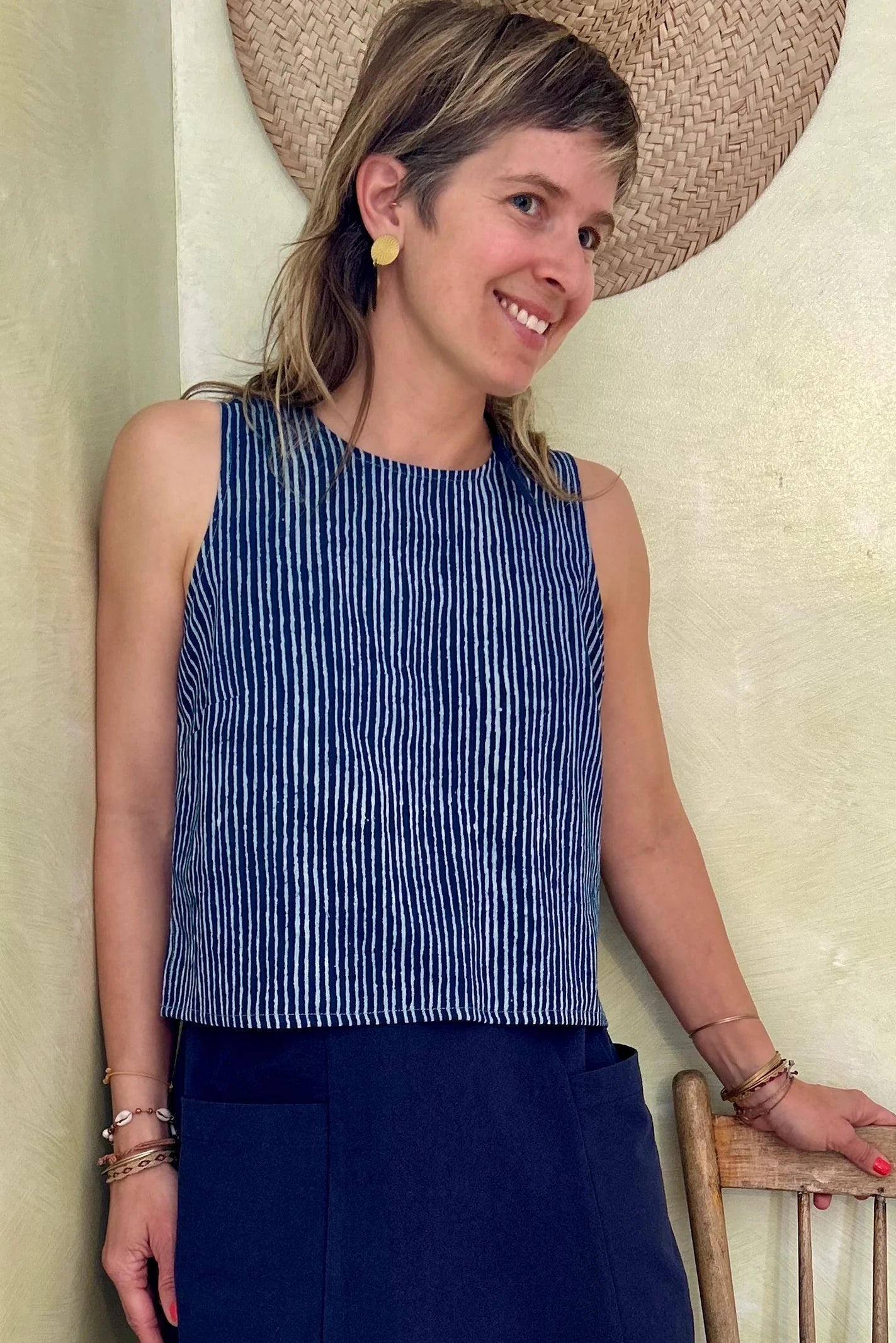 A woman wearing Forsythia top from Kazak in Indigo stripe, a cropped tank with a cutout and tie detail at the back. She is wearing it with a navy skirt and standing against a light-coloured wall. 