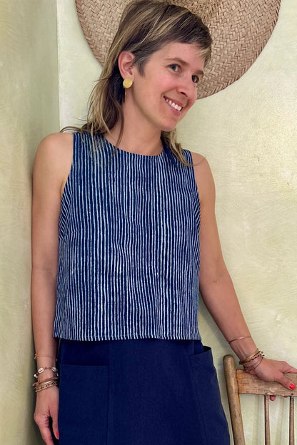 A woman wearing Forsythia top from Kazak in Indigo stripe, a cropped tank with a cutout and tie detail at the back. She is wearing it with a navy skirt and standing against a light-coloured wall. 