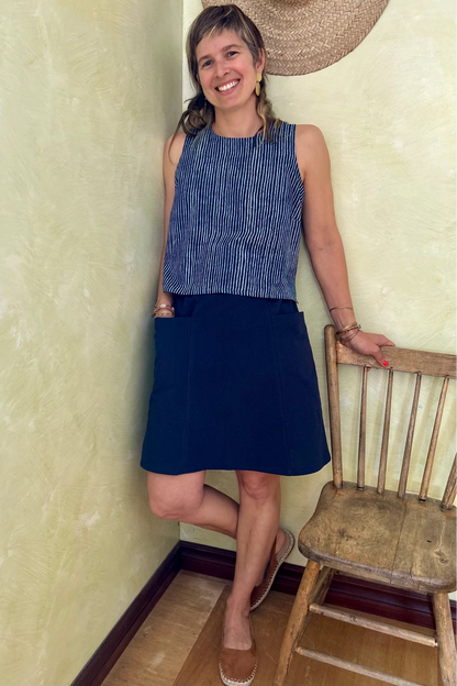 A woman wearing Forsythia top from Kazak in Indigo stripe, a cropped tank with a cutout and tie detail at the back. She is wearing it with a navy skirt and standing beside a wooden chair.  