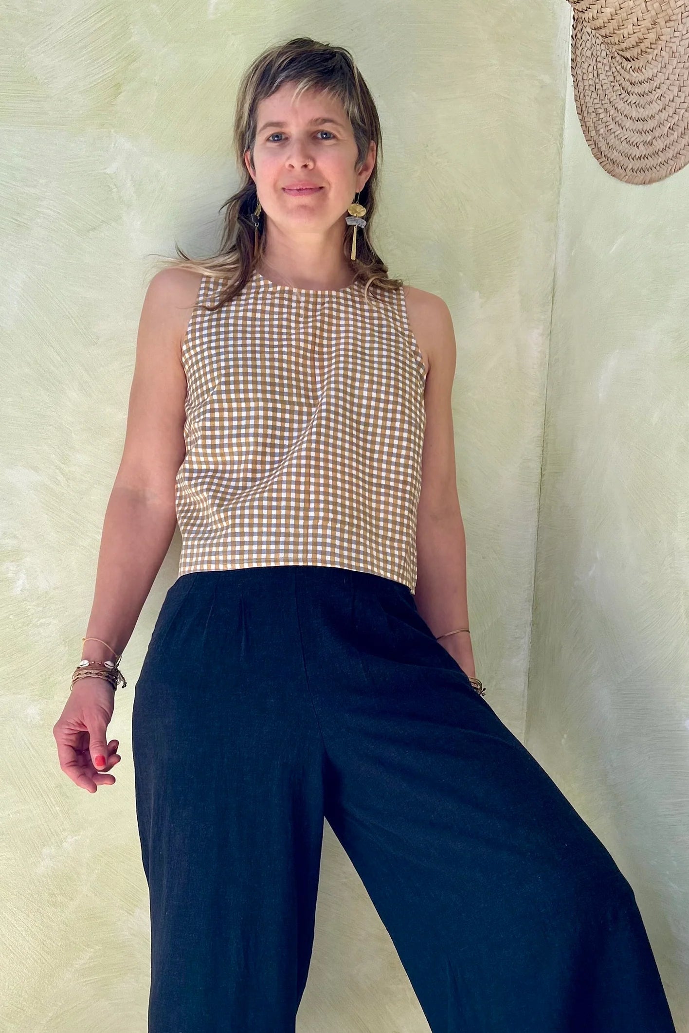 A woman wearing Forsythia top from Kazak in Mustard Vichy/Gingham,  a cropped tank with a cutout and tie detail at the back. She is wearing it navy pants and standing in front of a beige wall. 