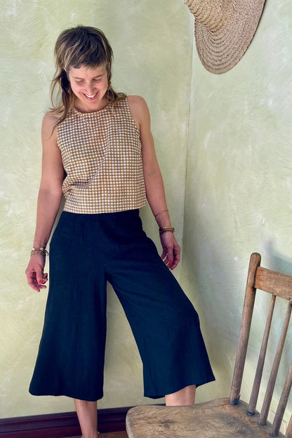 A woman wearing Forsythia top from Kazak in Mustard Vichy/Gingham,  a cropped tank with a cutout and tie detail at the back. She is wearing it navy pants and standing in front of a beige wall. 