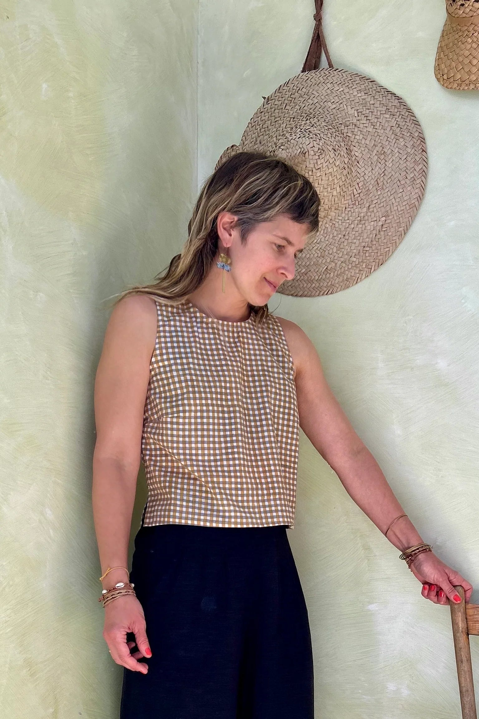 A woman wearing Forsythia top from Kazak in Mustard Vichy/Gingham,  a cropped tank with a cutout and tie detail at the back. She is wearing it navy pants and standing in front of a beige wall. 