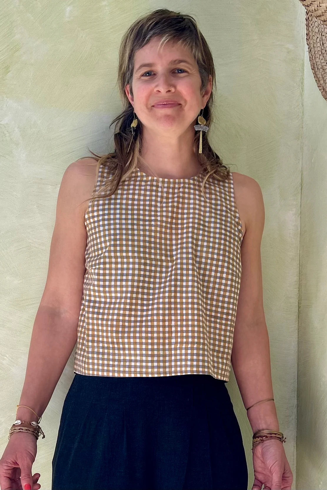 A woman wearing Forsythia top from Kazak in Mustard Vichy/Gingham,  a cropped tank with a cutout and tie detail at the back. She is wearing it navy pants and standing in front of a beige wall. 