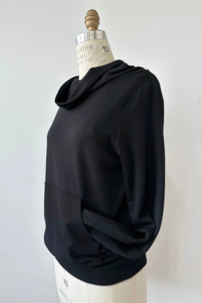 The Jullian Sweater by Kollontai in Black, with a cowl neck, long sleeves, and a kangaroo pocket, is shown on a mannequin in front of a white background. 
