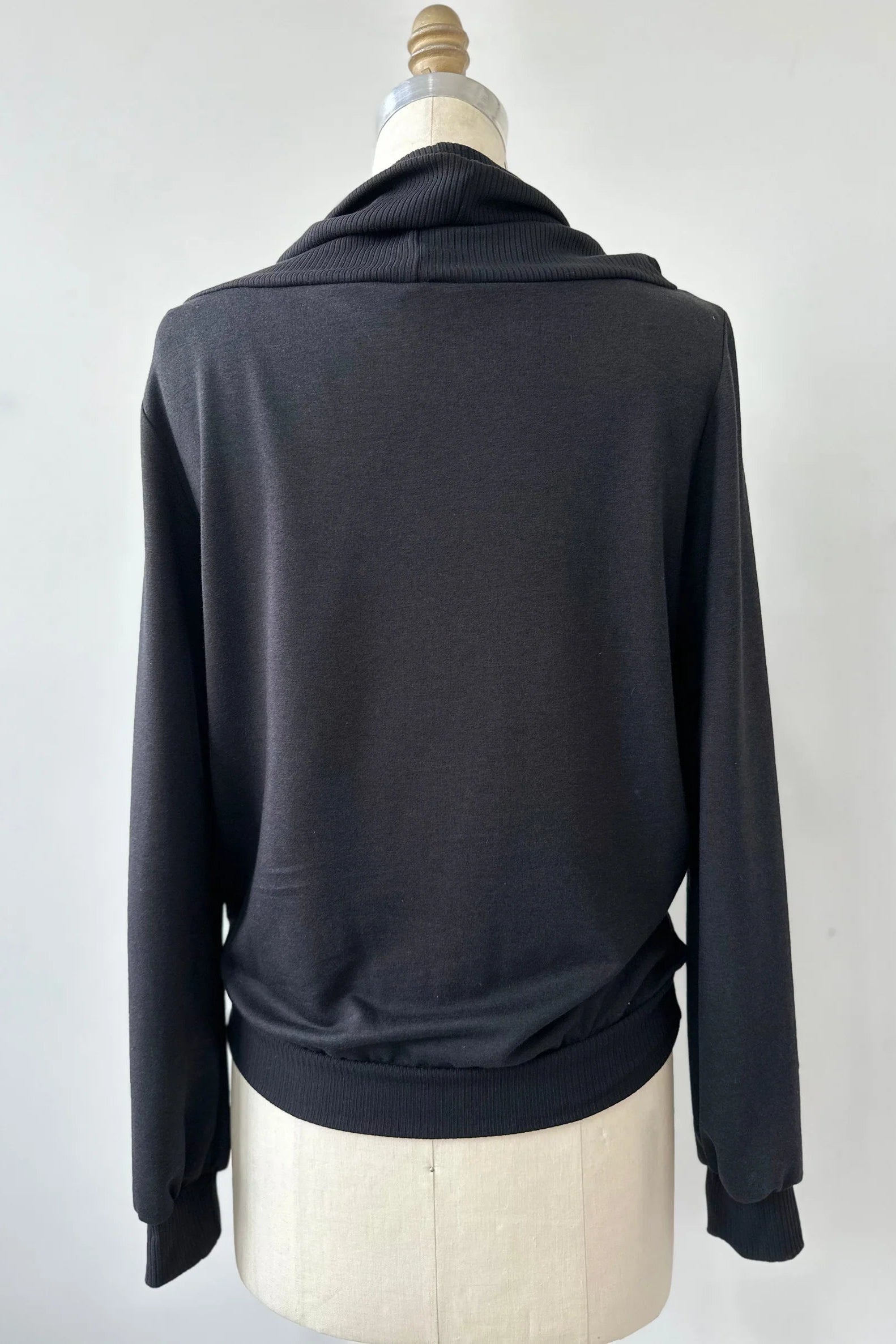 A back view of the Jullian Sweater by Kollontai in Black, with a cowl neck, long sleeves, and a kangaroo pocket, is shown on a mannequin in front of a white background. 