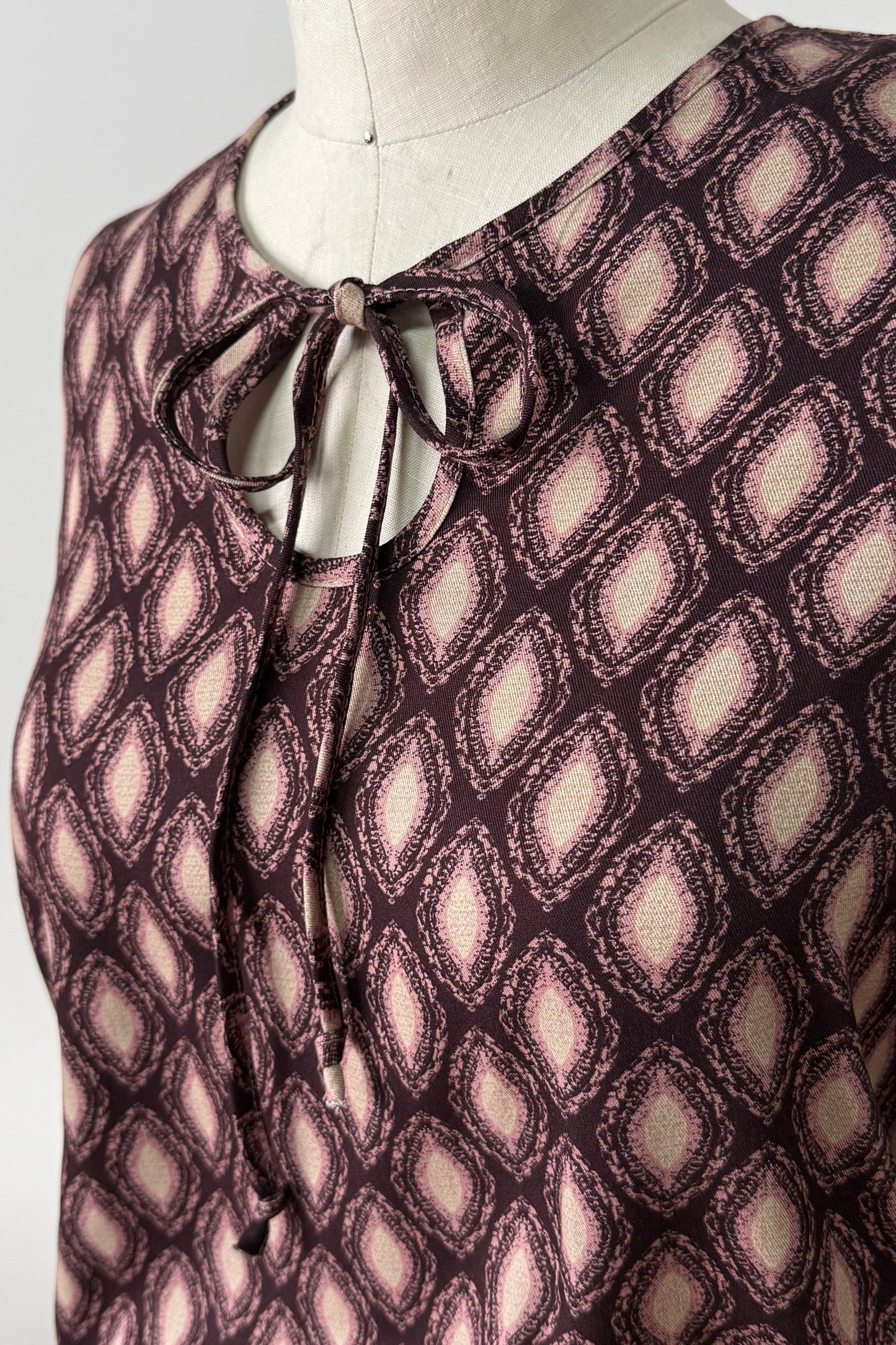 A close up of the Isabella Blouse by Kollontai, a burgundy and pink patterned blouse with a tie detail at the neck, balloon sleeves with square jewel buttons, and an elastic waist, is shown on a mannequin against a white background. 