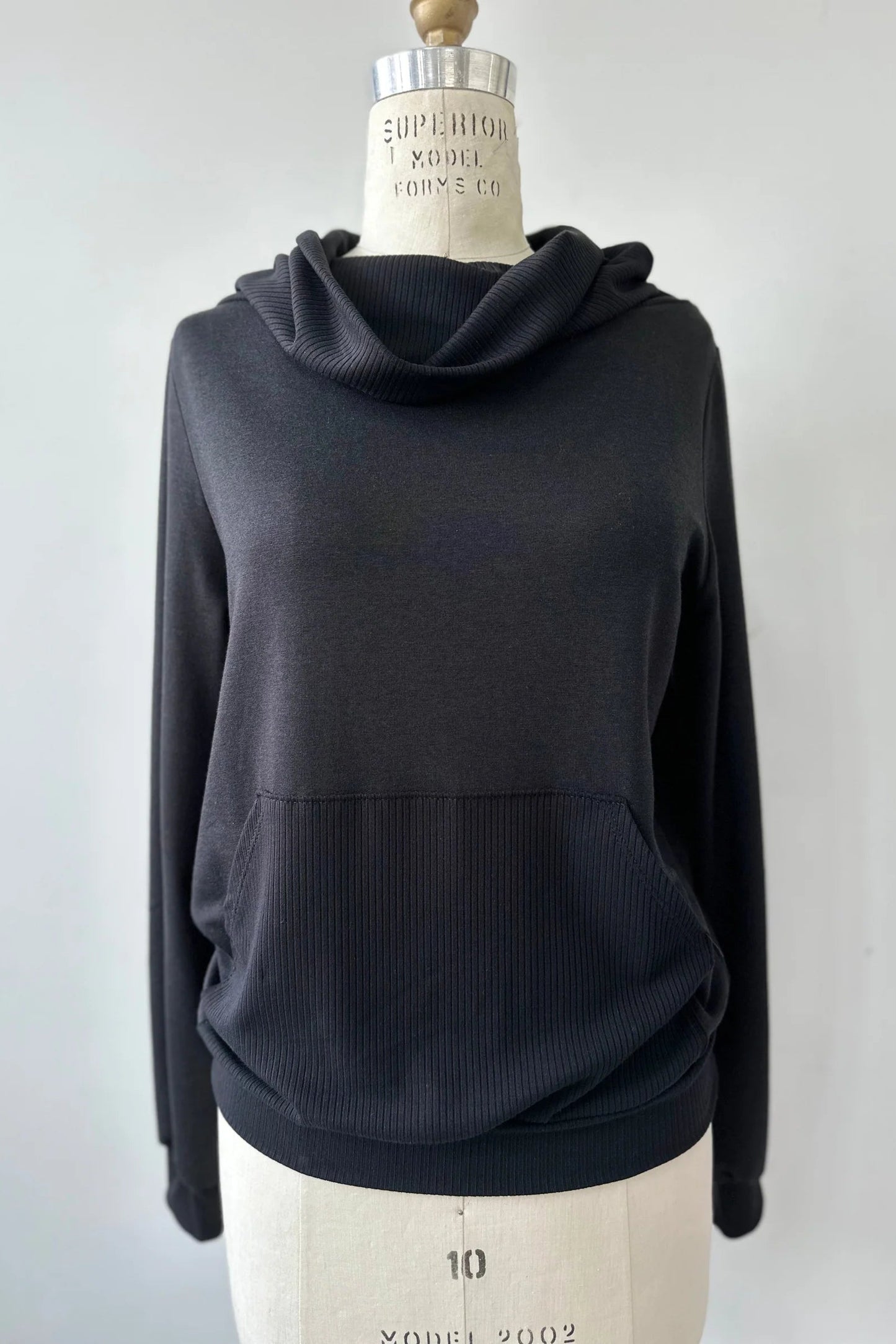 The Jullian Sweater by Kollontai in Black, with a cowl neck, long sleeves, and a kangaroo pocket, is shown on a mannequin in front of a white background. 