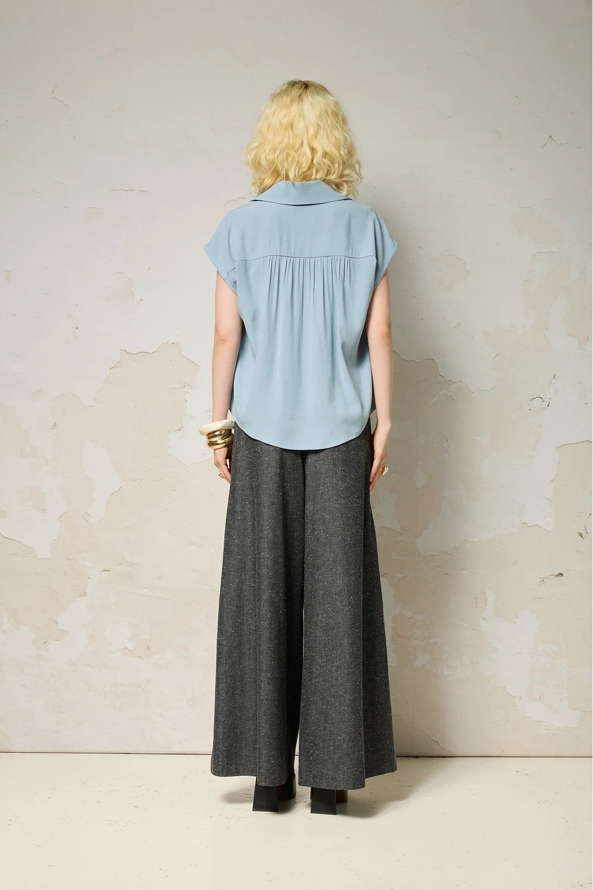 Back view of a woman wearing the Hemera Shirt by Cokluch in Sky, with a V-neck collar, button front, short extended sleeves, and a relaxed fit. The hem is hip-length in the front and longer at the back. She's wearing it with denim pants and standing in front of a grey background. 
