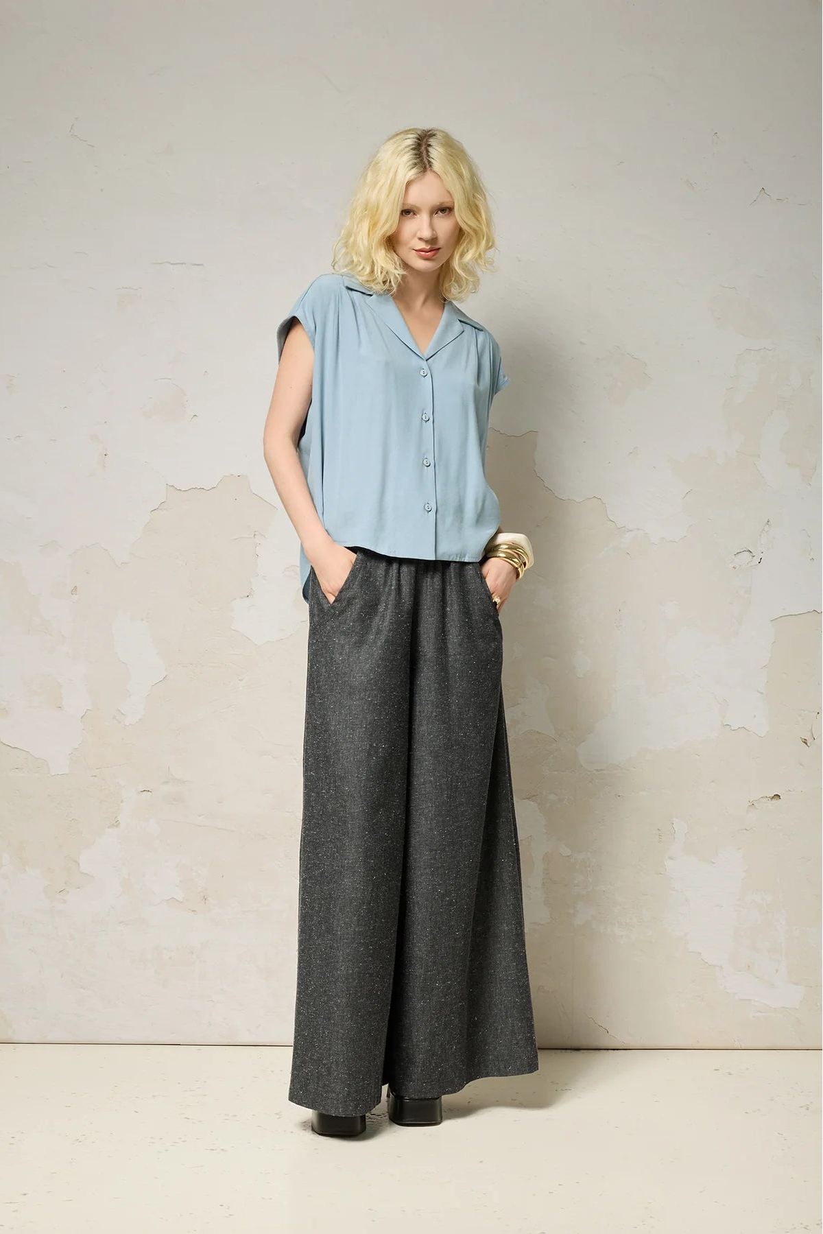 A woman wearing the Hemera Shirt by Cokluch in Sky, with a V-neck collar, button front, short extended sleeves, and a relaxed fit. The hem is hip-length in the front and longer at the back. She's wearing it with denim pants and standing in front of a grey background. 