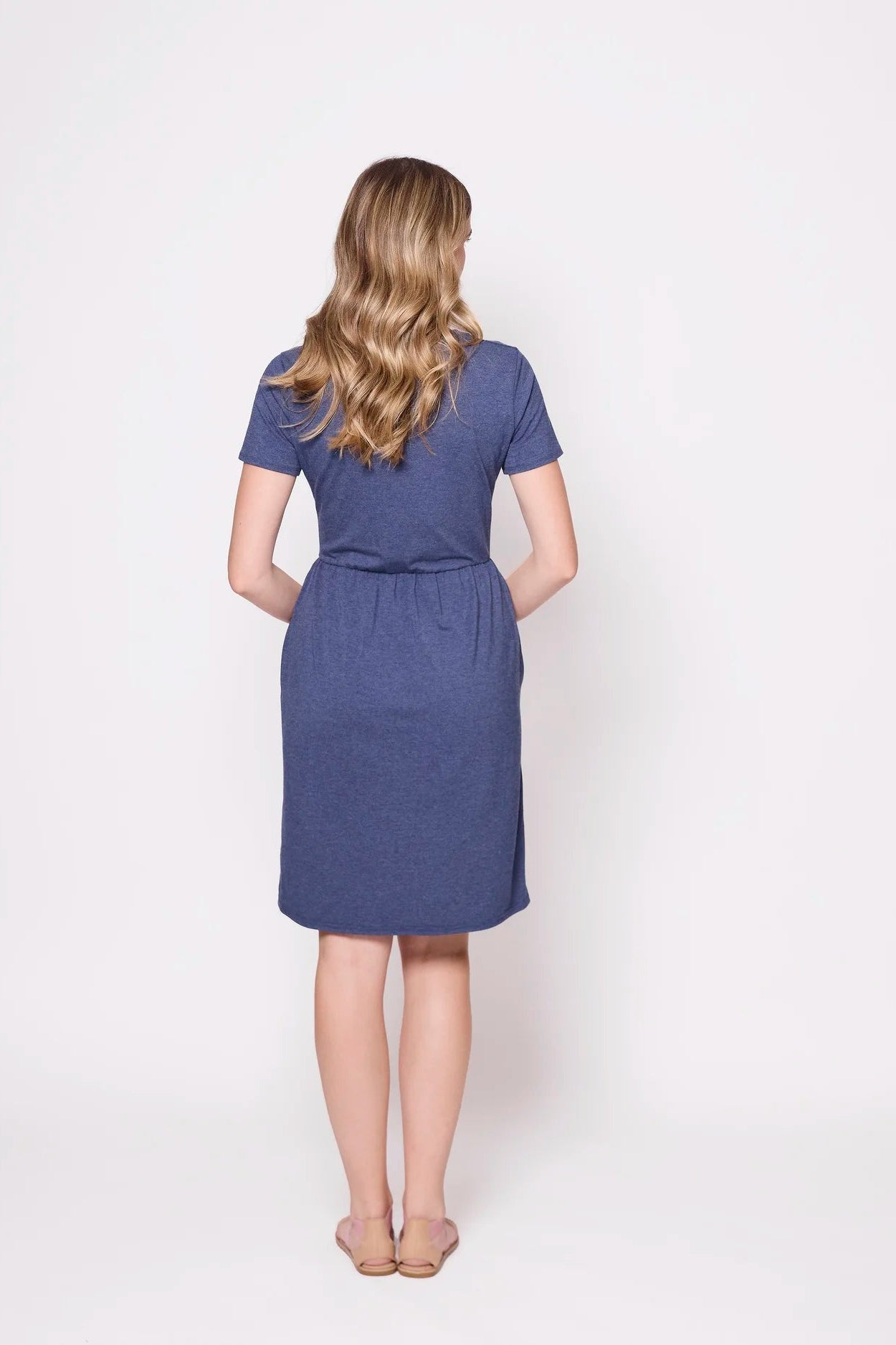 Back view of a woman wearing the Hayley Dress by Tangente in Heather Lake, a short sleeved bamboo dress with a gathered knee-length skirt. She is standing in front of a white background.
