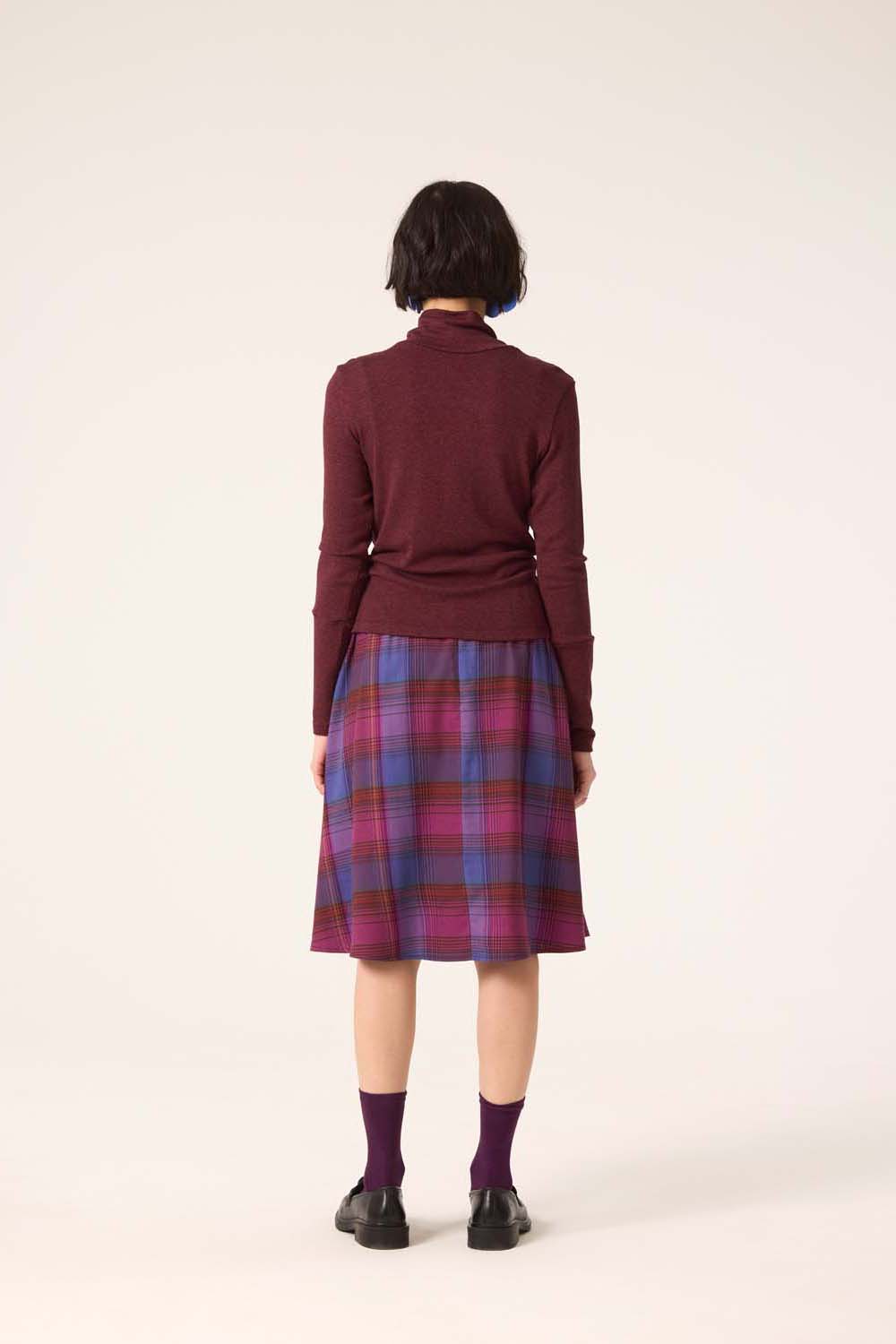 A woman wearing the Heritage Turtleneck by Cokluch in Shiraz with a plaid skirt, standing in front of a white background