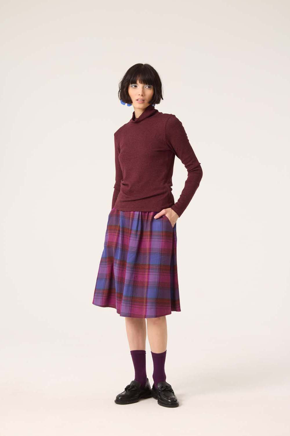 A woman wearing the Heritage Turtleneck by Cokluch in Shiraz with a plaid skirt, standing in front of a white background