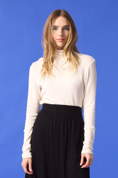 3/4 shot of a woman wearing the Heritage Turtleneck by Cokluch in Ivory with a dark skirt, standing in front of a blue background