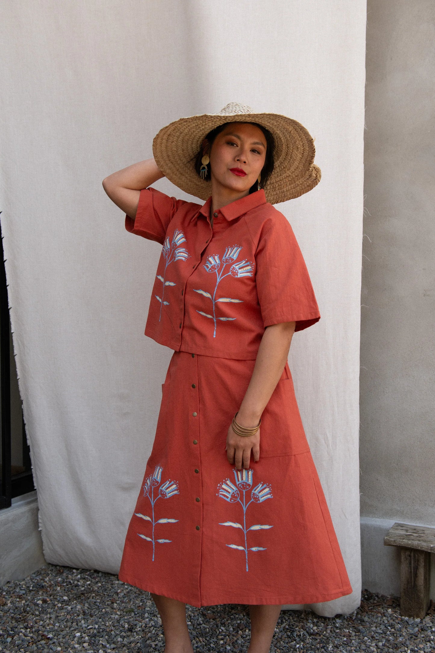 A woman wearing the Hosta Screen-printed Blouse by Kazak in Red with Pysanka, with hand-screen-printed flowers, mid-length raglan sleeves, a classic collar, and a button front. She is wearing it with a matching skirt and straw hat and standing outside by white curtain.
