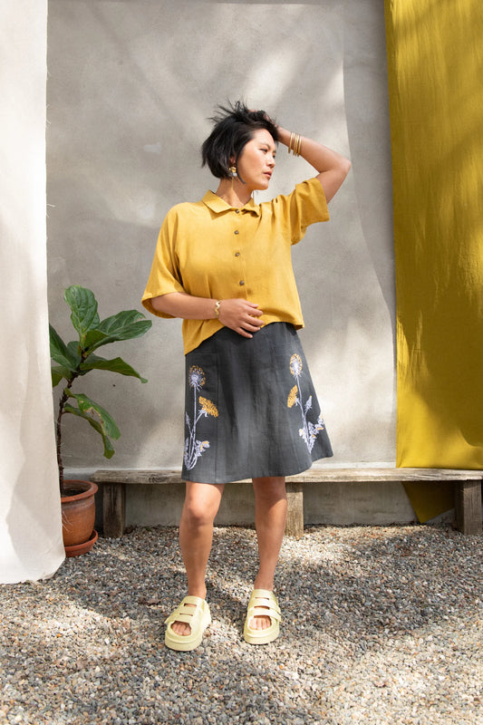 A woman wearing the Vicenze Screen-printed Skirt by Kazak in Black with Dandelions, an above the knee A-line skirt with hand-screened flowers, an elasticated waist, and large patch pockets. She is wearing it with the  Hosta shirt in Mustard and standing outside by a grey wall and white and yellow curtains.