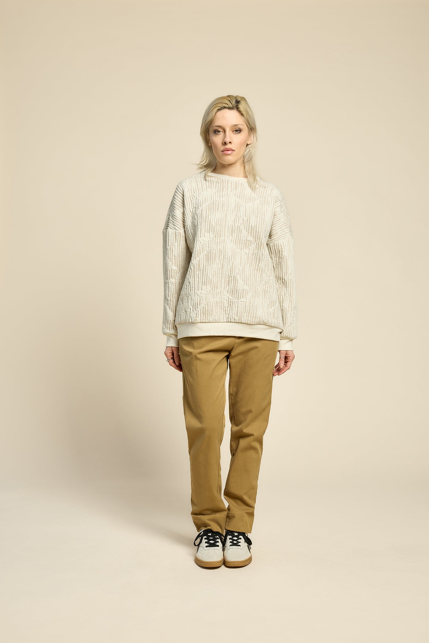 A woman wearing the Hotel Sweater by Cokluch in Natural, a loose fitting round necked sweater in Crinkle Max fabric. She is wearing it with yellow pants and standing in front of a beige background. 