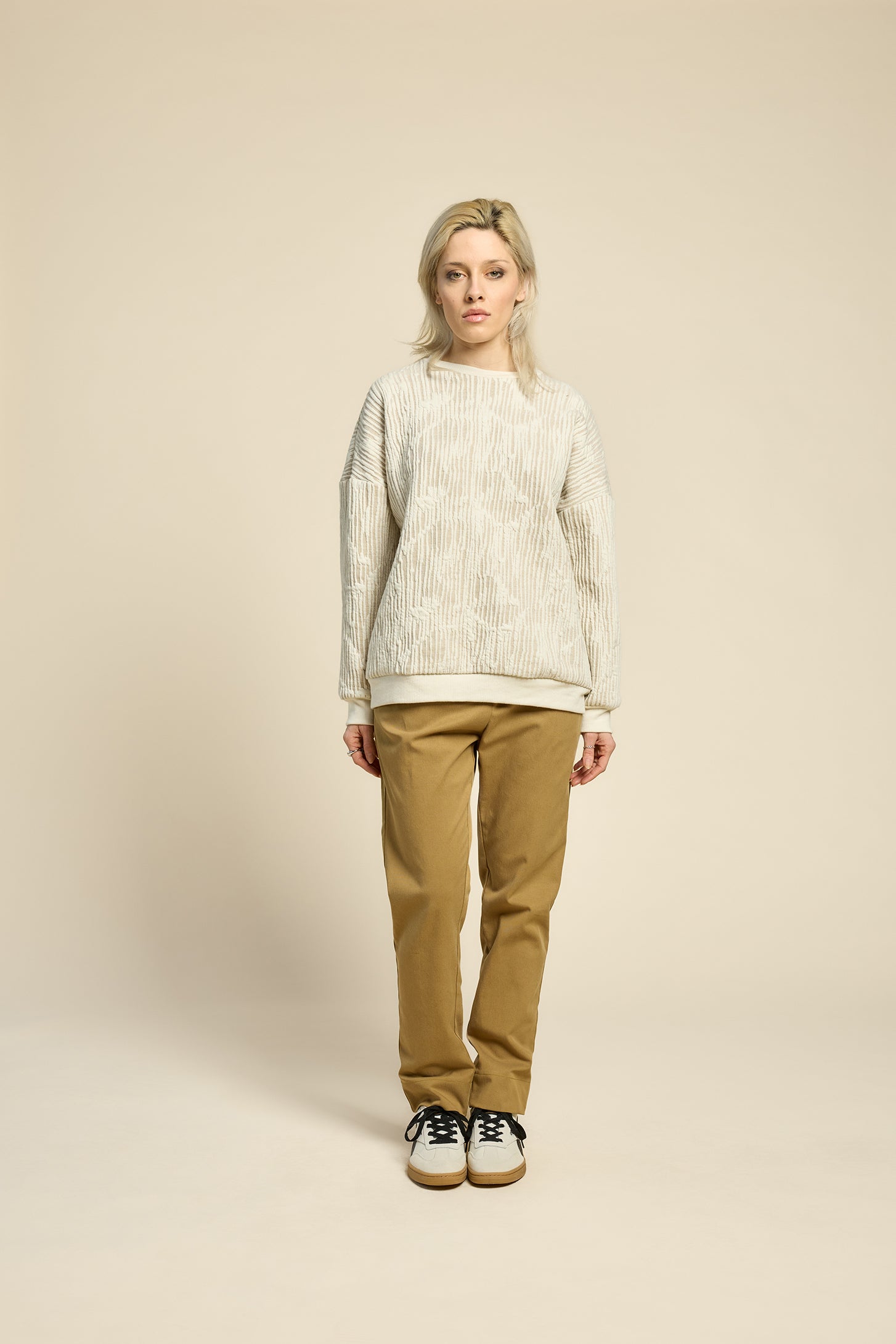 A woman wearing the Hotel Sweater by Cokluch in Natural, a loose fitting round necked sweater in Crinkle Max fabric. She is wearing it with yellow pants and standing in front of a beige background. 