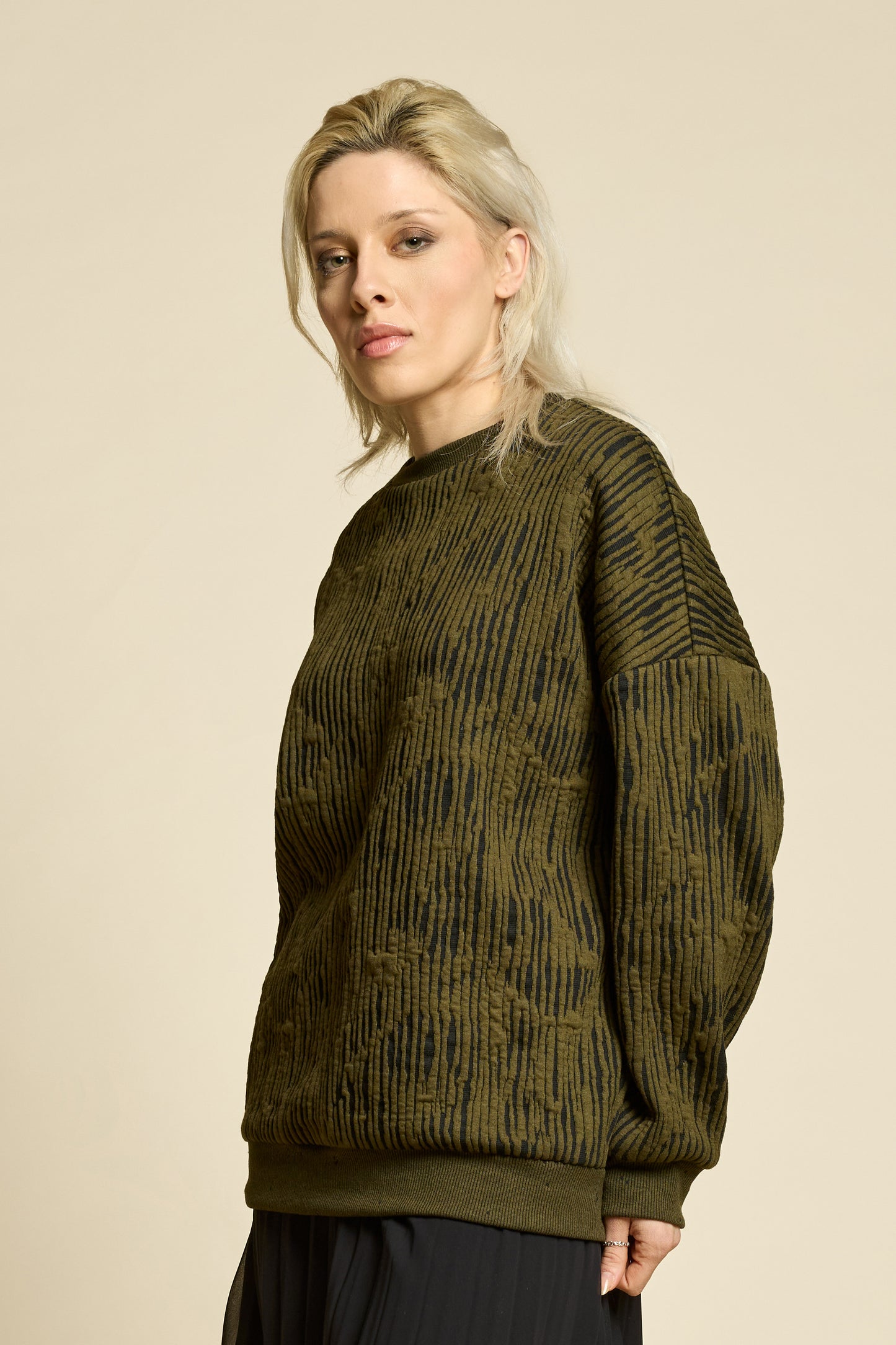 A woman wearing the Hotel Sweater by Cokluch in Olive, a loose fitting round necked sweater in Crinkle Max fabric. She is wearing it with black pants and standing in front of a beige background. 