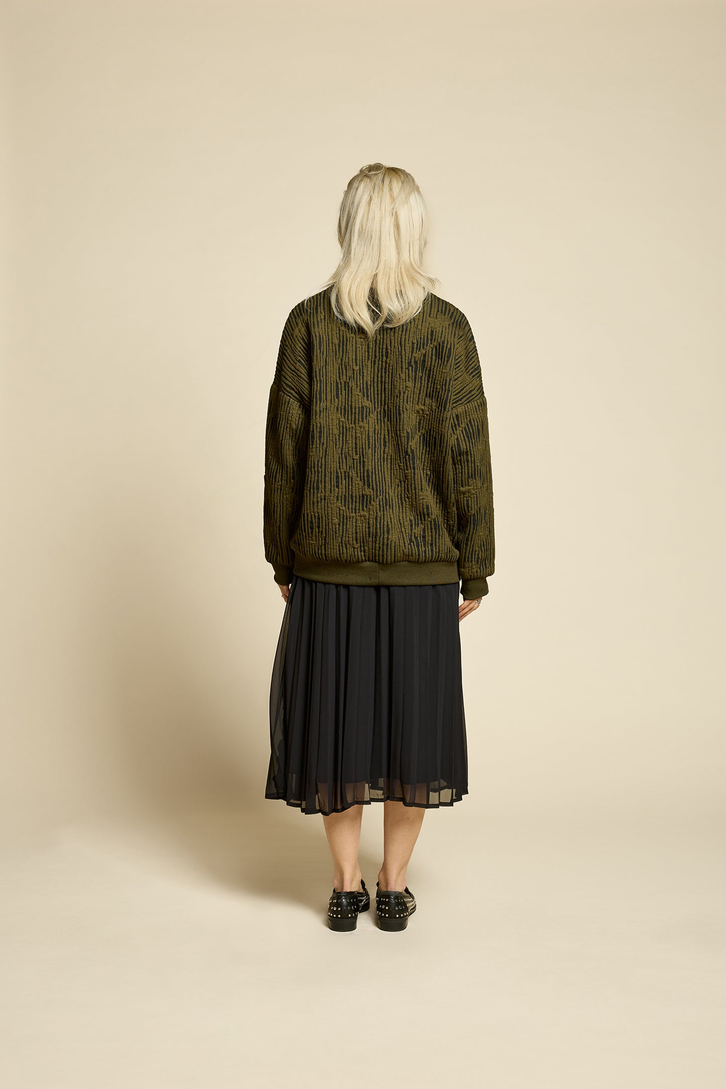 Back view of a woman wearing the Hotel Sweater by Cokluch in Olive, a loose fitting round necked sweater in Crinkle Max fabric. She is wearing it with a bladck skirt and standing in front of a beige background. 