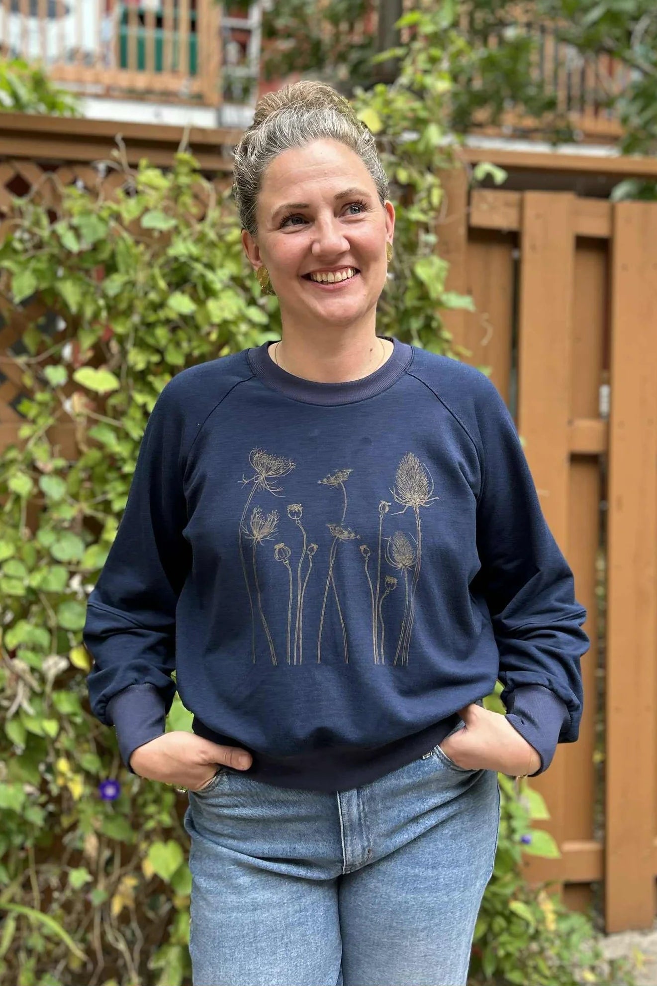 A woman wearing the Marilou Crewneck with Flowers by Kazak in Navy, a raglan sleeved sweatshirt with a matte bronze screen print design. She is wearing it with jeans and standing in a garden. 