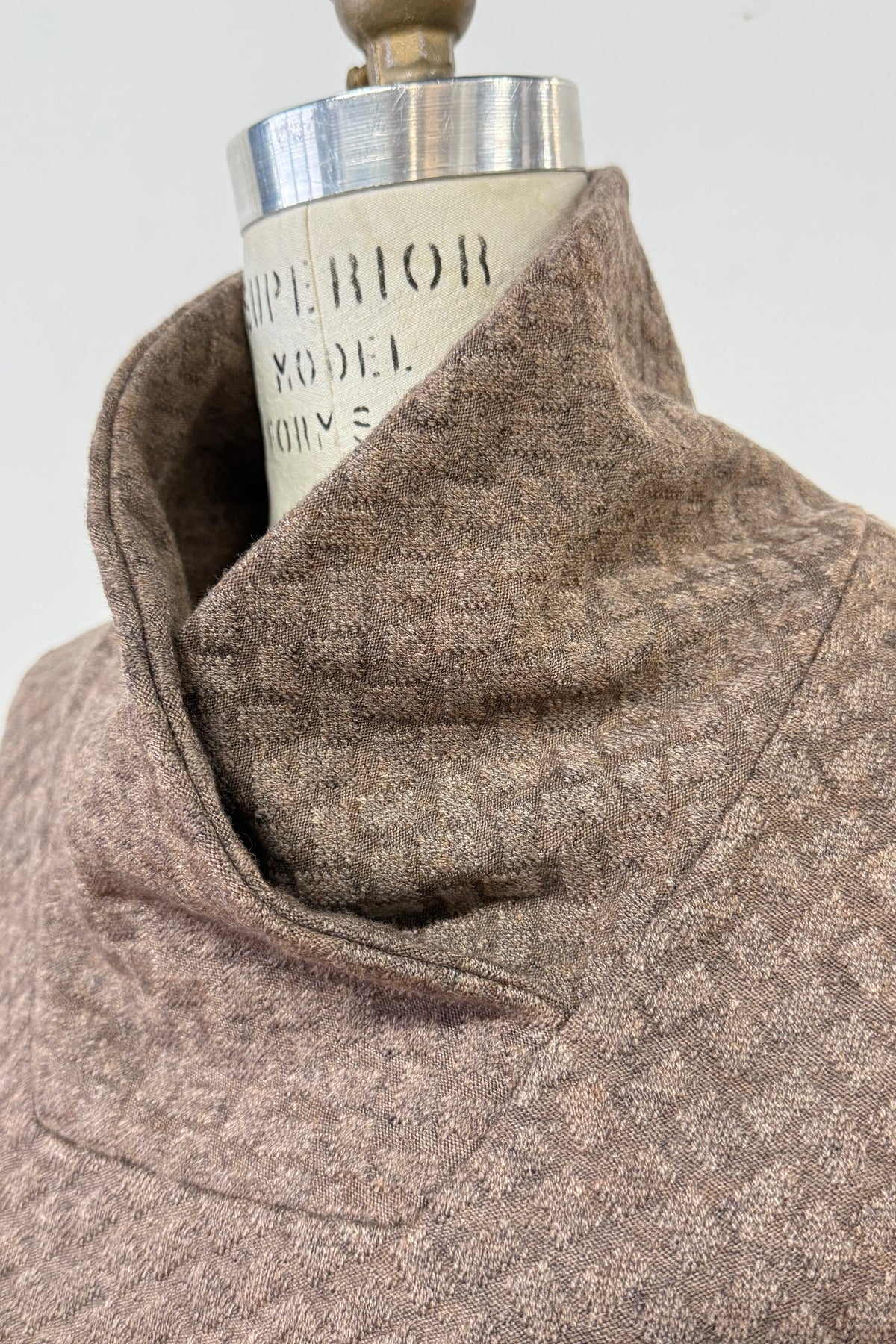A close up of the collar of the Sacha Tunic by Kollontai in Brown, a textured knit straight cut tunic with a funnel collar and large patch pockets, is shown on a mannequin against a white background. 