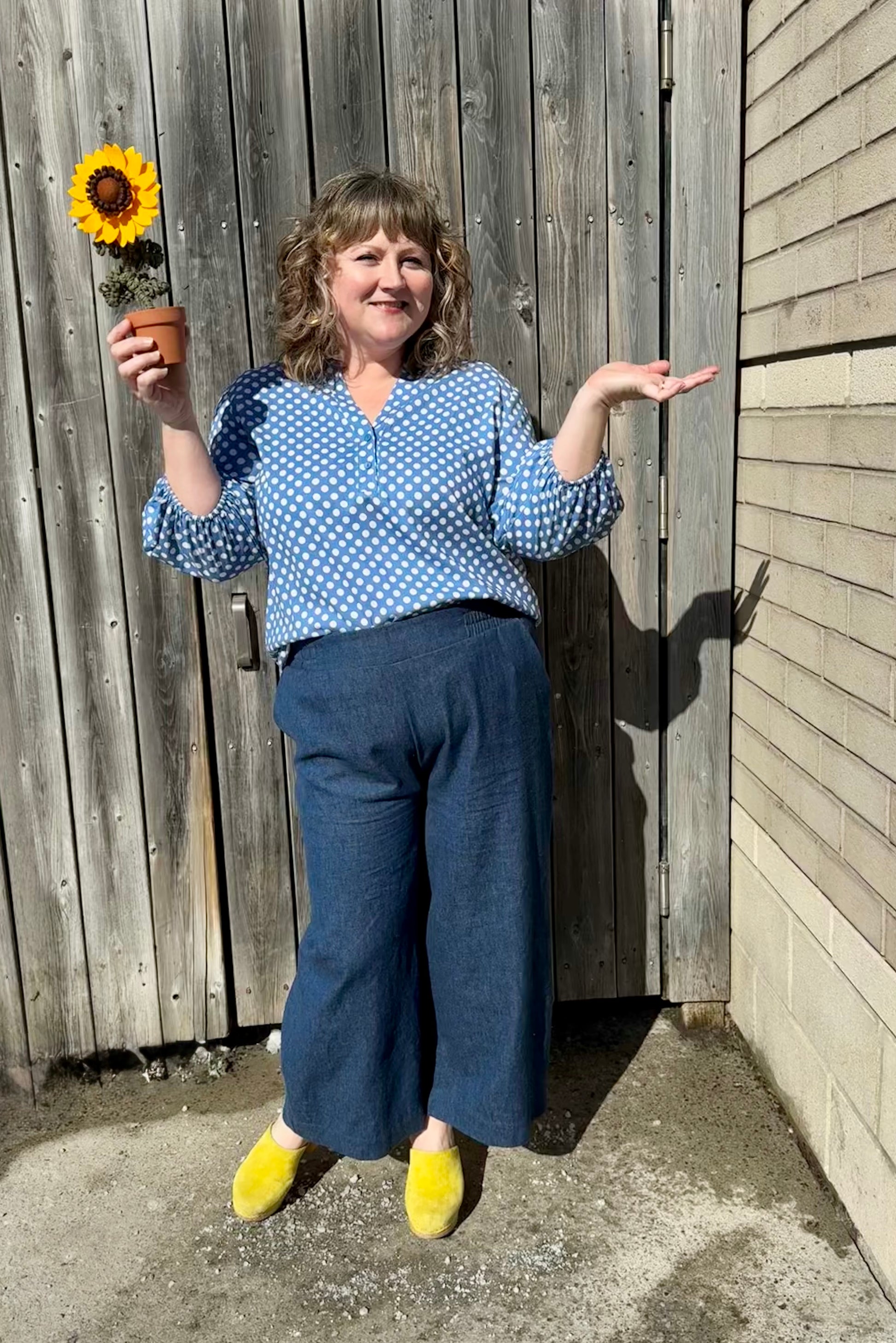 A woman wearing the Grace Blouse by Pure Essence in Chambray Dot, with a v-placket neck with buttons and 7/8 gathered sleeves. She is wearing it with denim pants and standing against a wooden fence while holding a fabric sunflower. 
