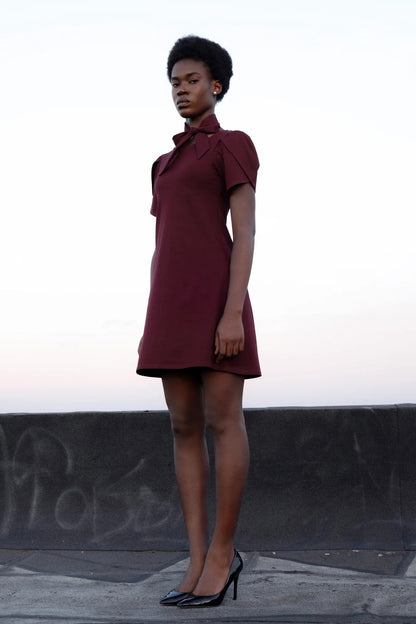 A woman wearing the Opal Dress by Eve Lavoie in burgundy, a short dress with tulip sleeves, an A-line shape, and a removable belt that's styled as a necktie. She's standing outdoors by a low concrete wall. 