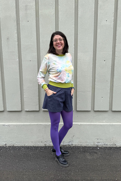 A woman wearing the Maya shorts from Cokluch in Indigo Speckle, lightweight denim shorts with a waist that's elasticized at the back, wide culotte style legs, and a side pockets. She is wearing them with the Wanda E26 Sweater by Slak in Watercolour print and purple tights and standing outside in front of panelled wall. 