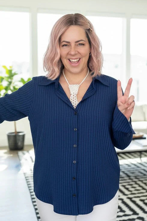 A woman wearing the Blue Striped Blouse by Dotty, a classic button front shirt with a collar and extra wide cuffs. She is wearing it with white pants and standing in a room with a plant and low table. 