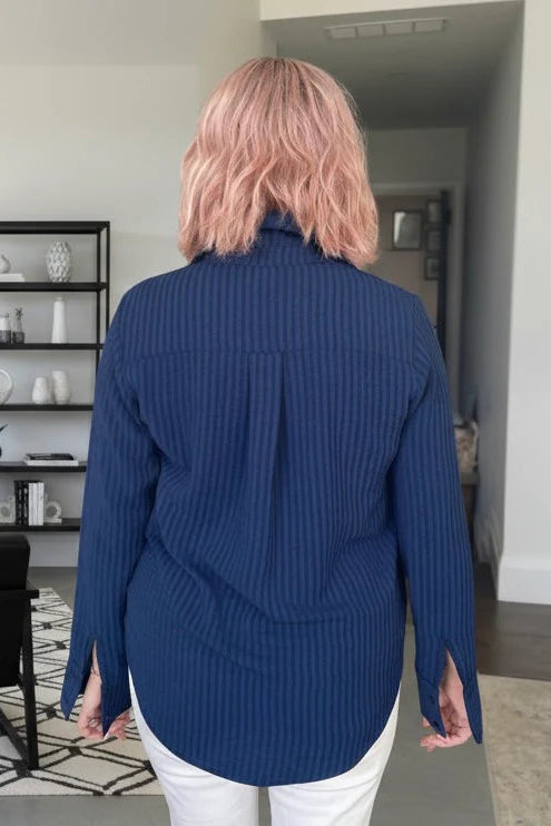 Back view of a woman wearing the Blue Striped Blouse by Dotty, a classic button front shirt with a collar and extra wide cuffs. She is wering it with white pants and standing in a room with shelves.