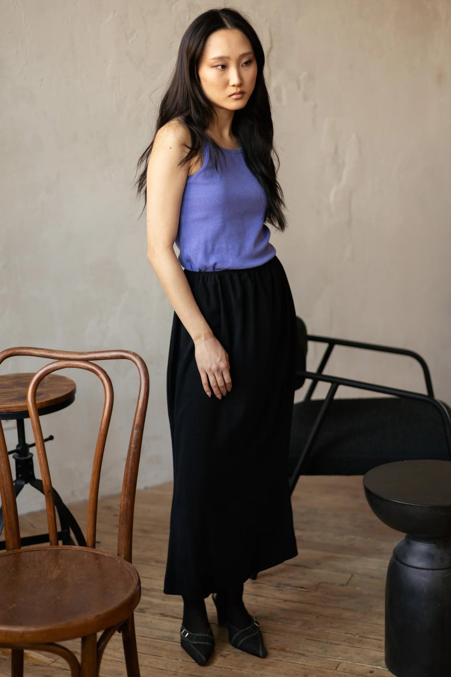 A woman wearing the Simone Skirt by Lights of All in Black, a sleek calf-length skirt with a flared hem and an elastic waist. She is wearing it with a purple top and standing in a room with furniture. 