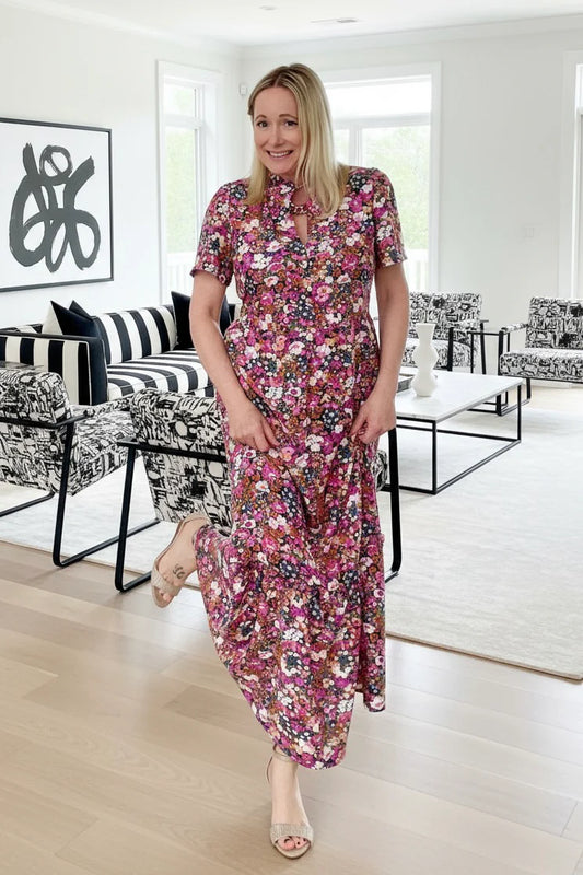 A woman wearing the Petal Print by Dotty in Pink/Blue floral,  a maxi dress with short flutter sleeves, a split V-neck and ruffled collar, with a gathered waist and A-line skirt. She is standing in a modern living room.
