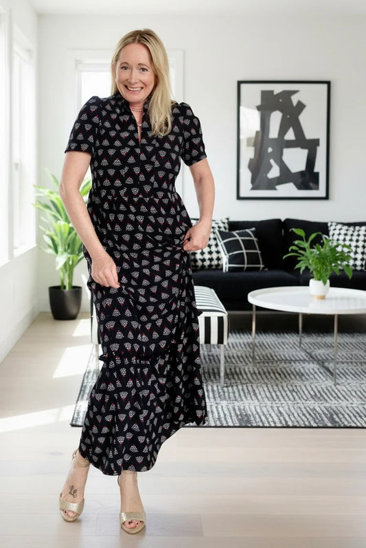 A woman wearing the Art Deco Dress by Dotty in Black print, a maxi dress with short flutter sleeves, a split V-neck and ruffled collar, with a gathered waist and A-line skirt. She is standing in a modern living room.