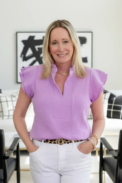 A woman wearing the Purple Cotton Blouse by Dotty, with short double ruffled sleeves, a split v-neck, ruffle-banded colour and rounded hi-low hem. She is wearing it with white pants in an indoor setting. 