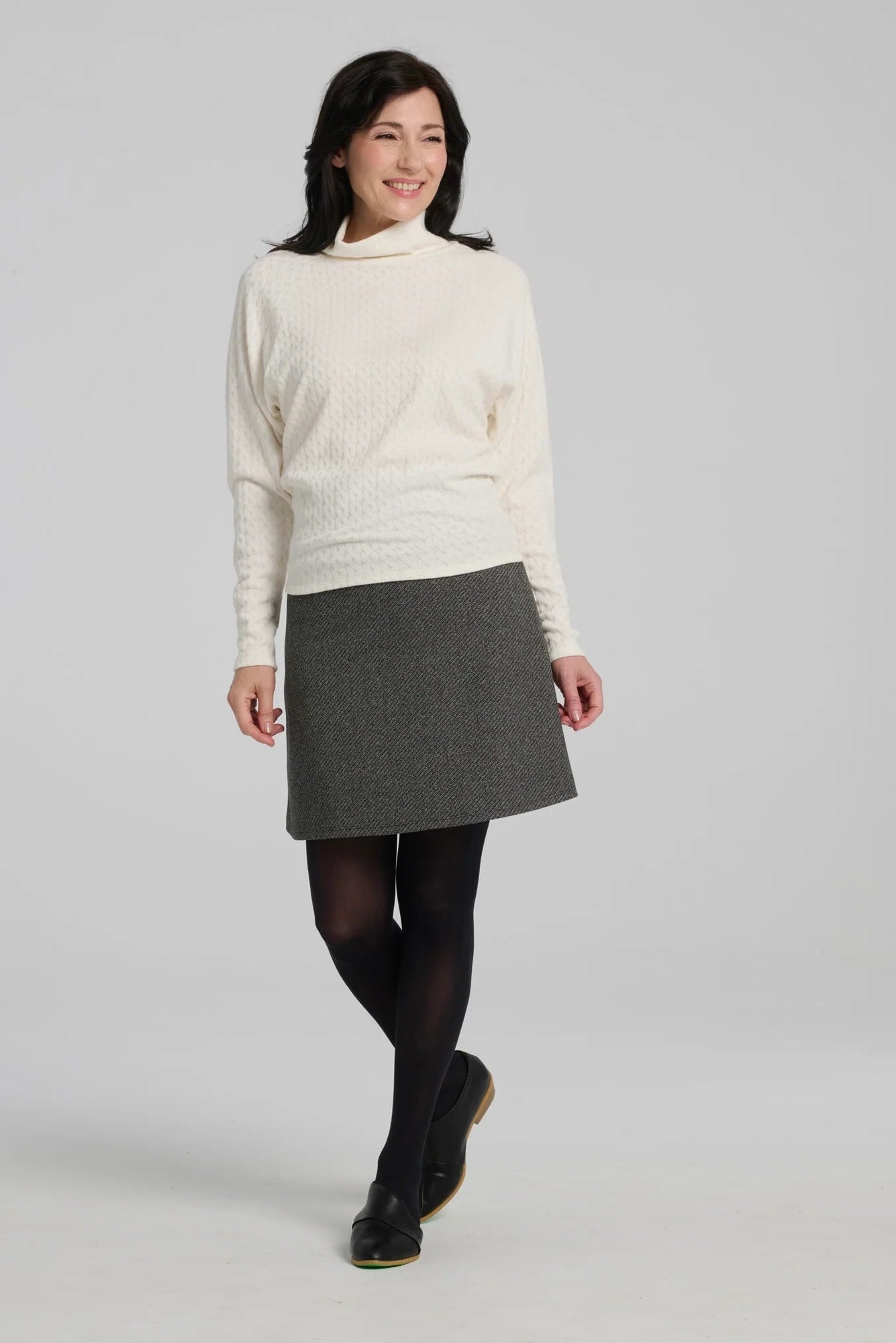A woman wearing the Plume Sweater by Kollontai in White, a cable knit turtleneck with long tapered sleeves and a narrow hem for a bloused look. She is wearing it with a grey skirt and standing in front of a grey background. 