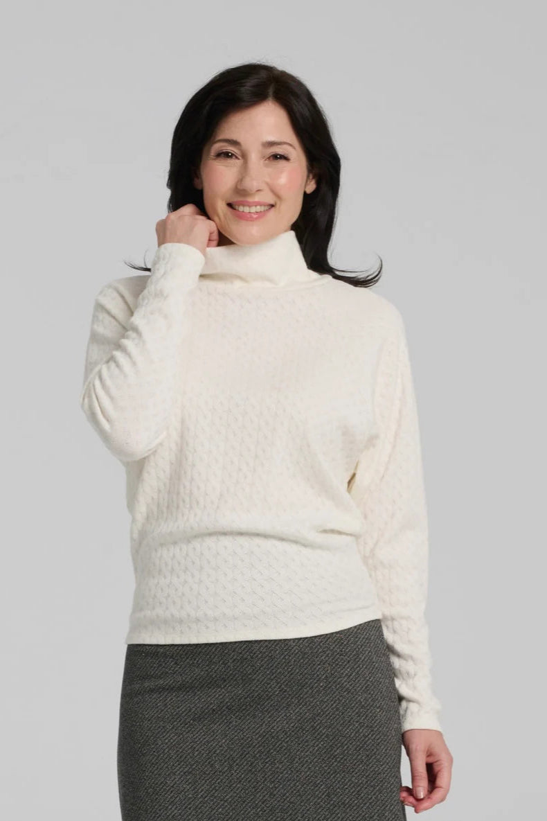 Waist up view of a woman wearing the Plume Sweater by Kollontai in White, a cable knit turtleneck with long tapered sleeves and a narrow hem for a bloused look. She is wearing it with a grey skirt and standing in front of a grey background. 
