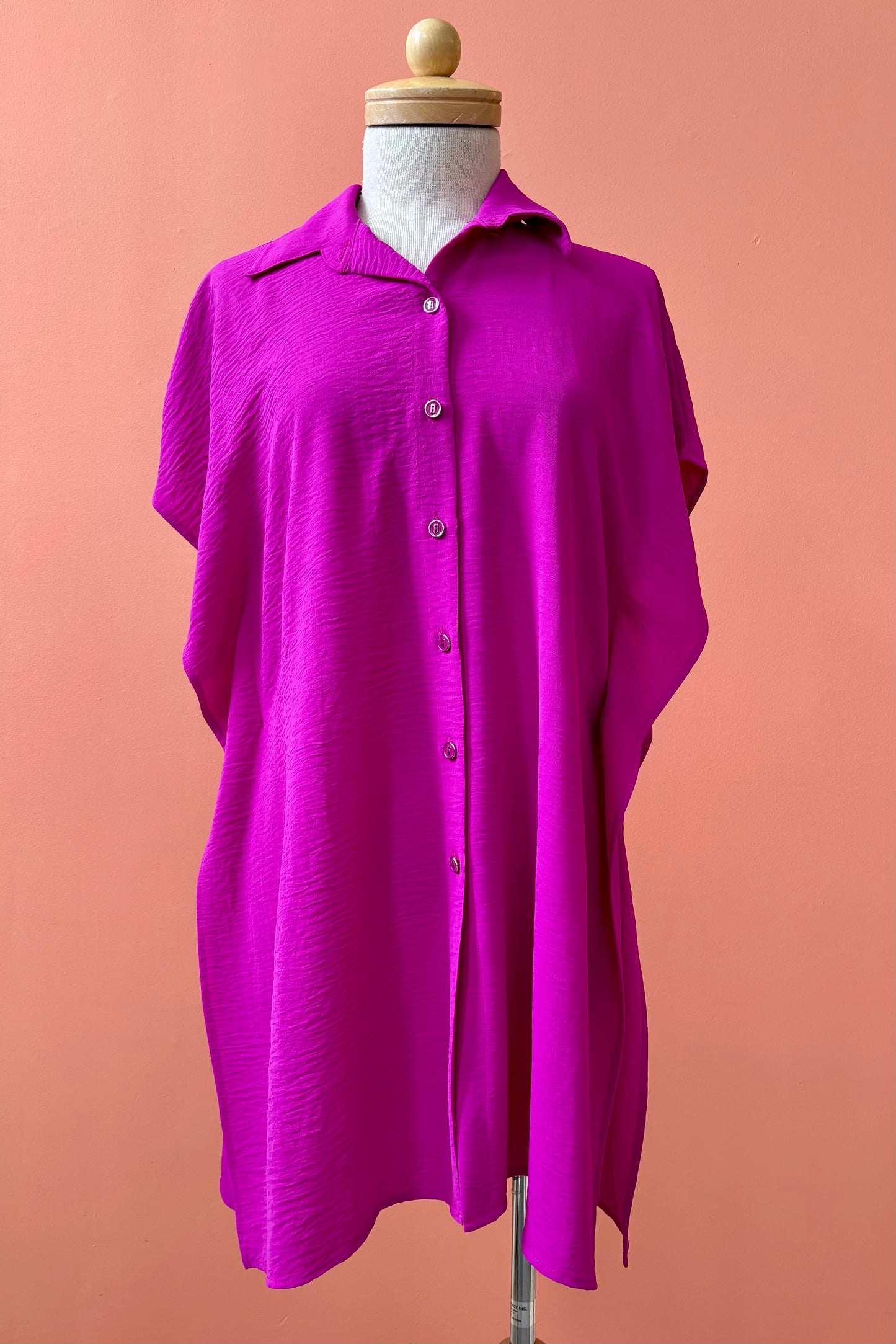 The Geneva Top by Pure Essence in Magenta, a long and loose button front shirt with short sleeves, a classic collar, and high side slits is shown on a mannequin against a coral background. 