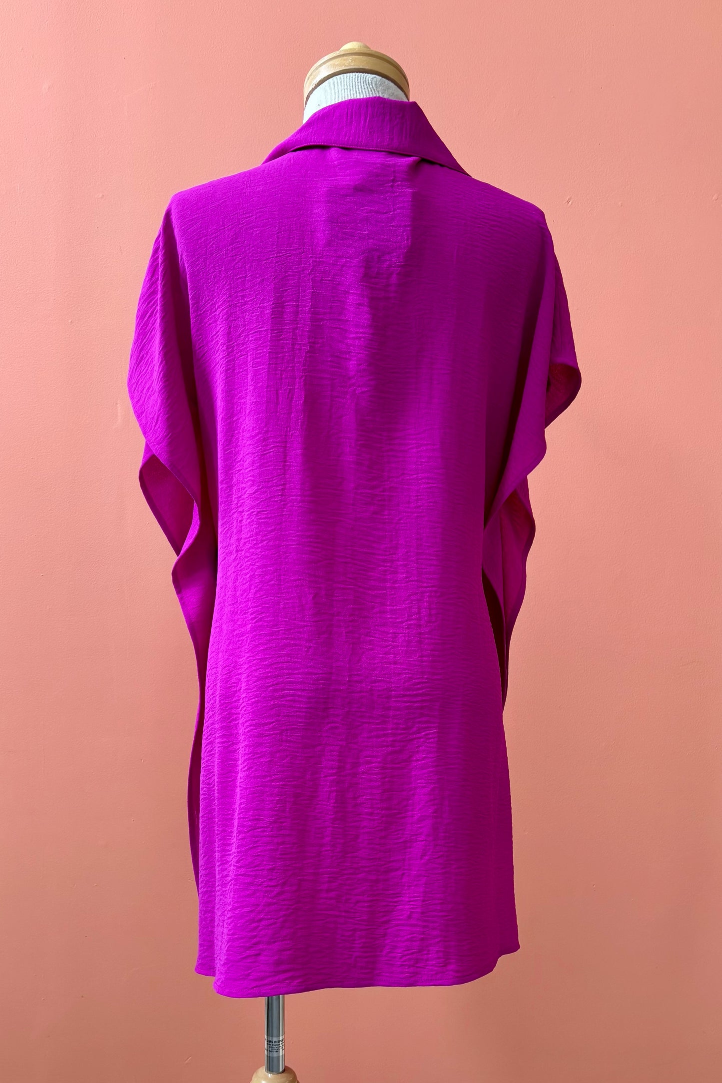 A back view of the Geneva Top by Pure Essence in Magenta, a long and loose button front shirt with short sleeves, a classic collar, and high side slits is shown on a mannequin against a coral background. 