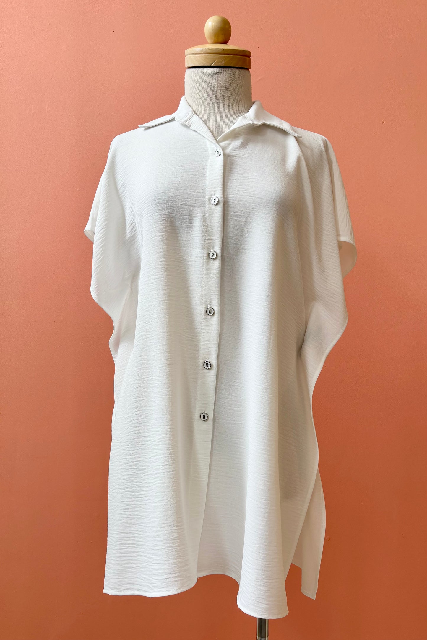 The Geneva Top by Pure Essence in White, a long and loose button front shirt with short sleeves, a classic collar, and high side slits is shown on a mannequin against a coral background. 