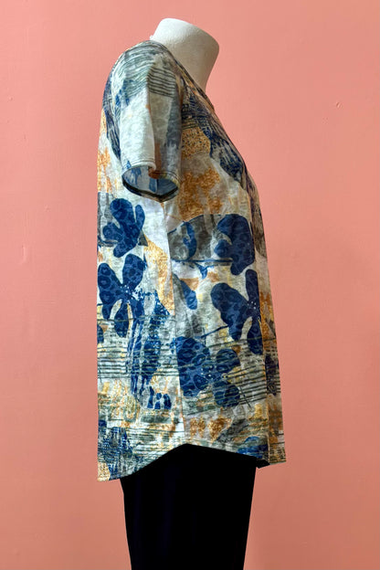 A side view of the Gladys T-shirt by Pure Essence in Golden Multi print is shown on a mannequin against a coral background. 