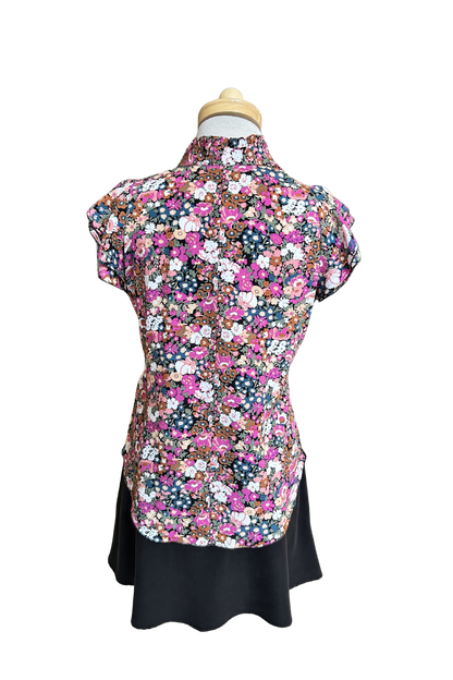 A back view of the Petal Blouse by Dotty in Pink/Blue floral print is shown on a mannequin against a white background. It has short floaty sleeves, a split V-neck, a ruffled collar, and a rounded hi-low hemline. 