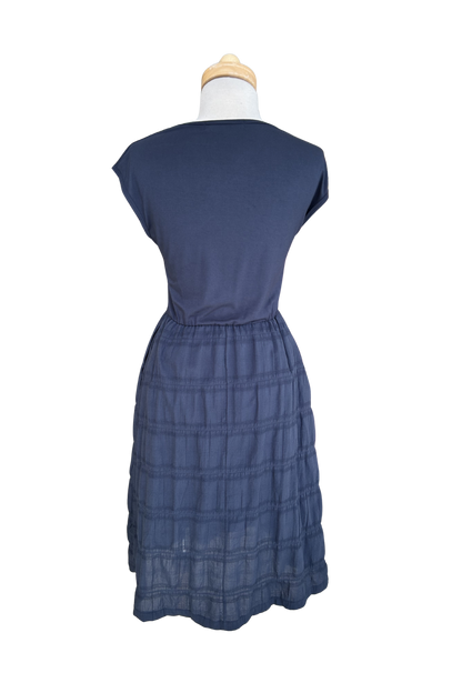 A back view of the Mona Dress by Tangente in Navy, a below the knee length dress featuring a bamboo rayon bodice and seersucker skirt, with short sleeves and a round neck, is shown on a mannequin against a white background. 
