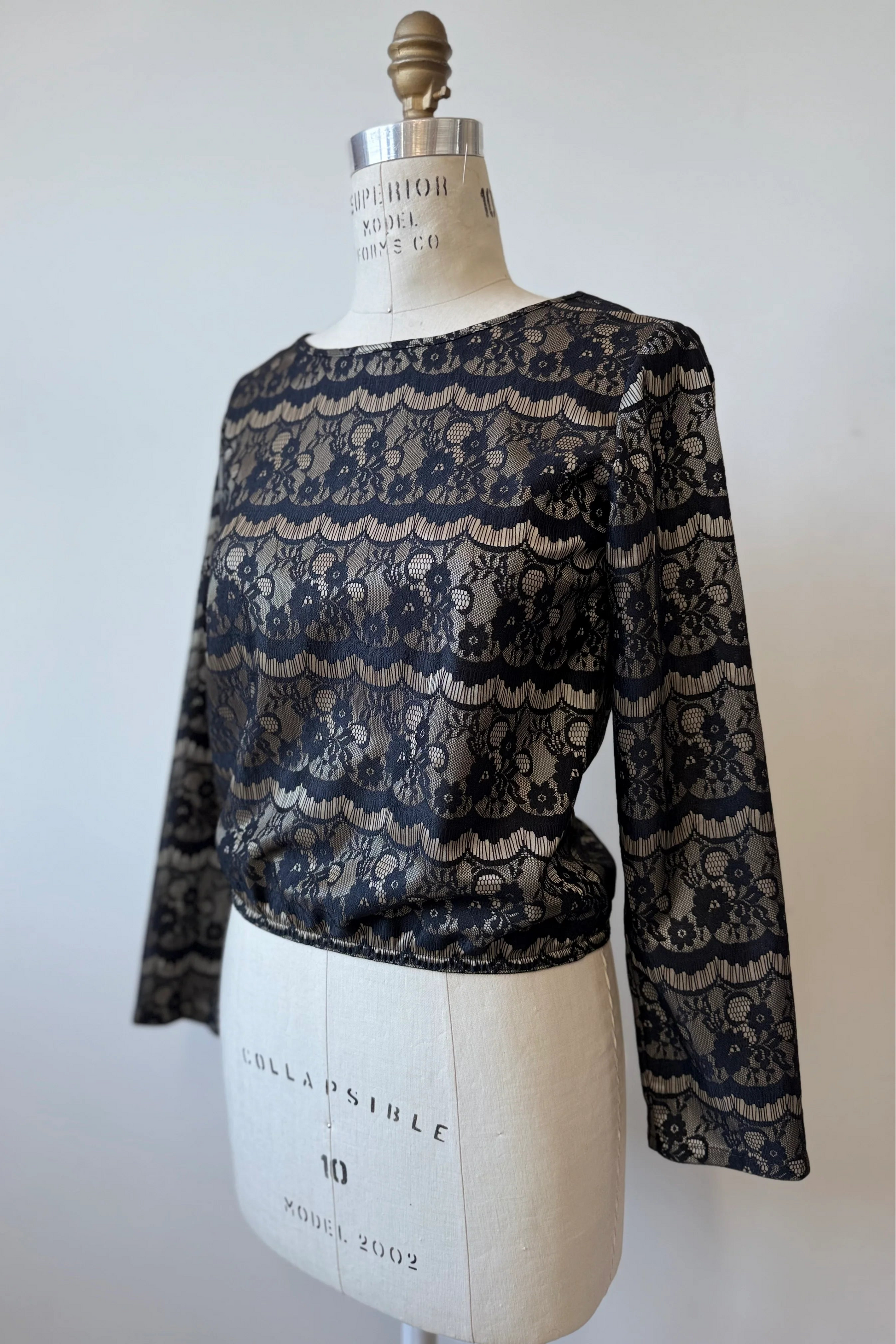 The Solstice Blouse by Kollontai in Gold is shown on a mannequin in front of a white background.  The fabric layers black lace over golden satin. It's slightly cropped with an elastic waist and long sleeves. 