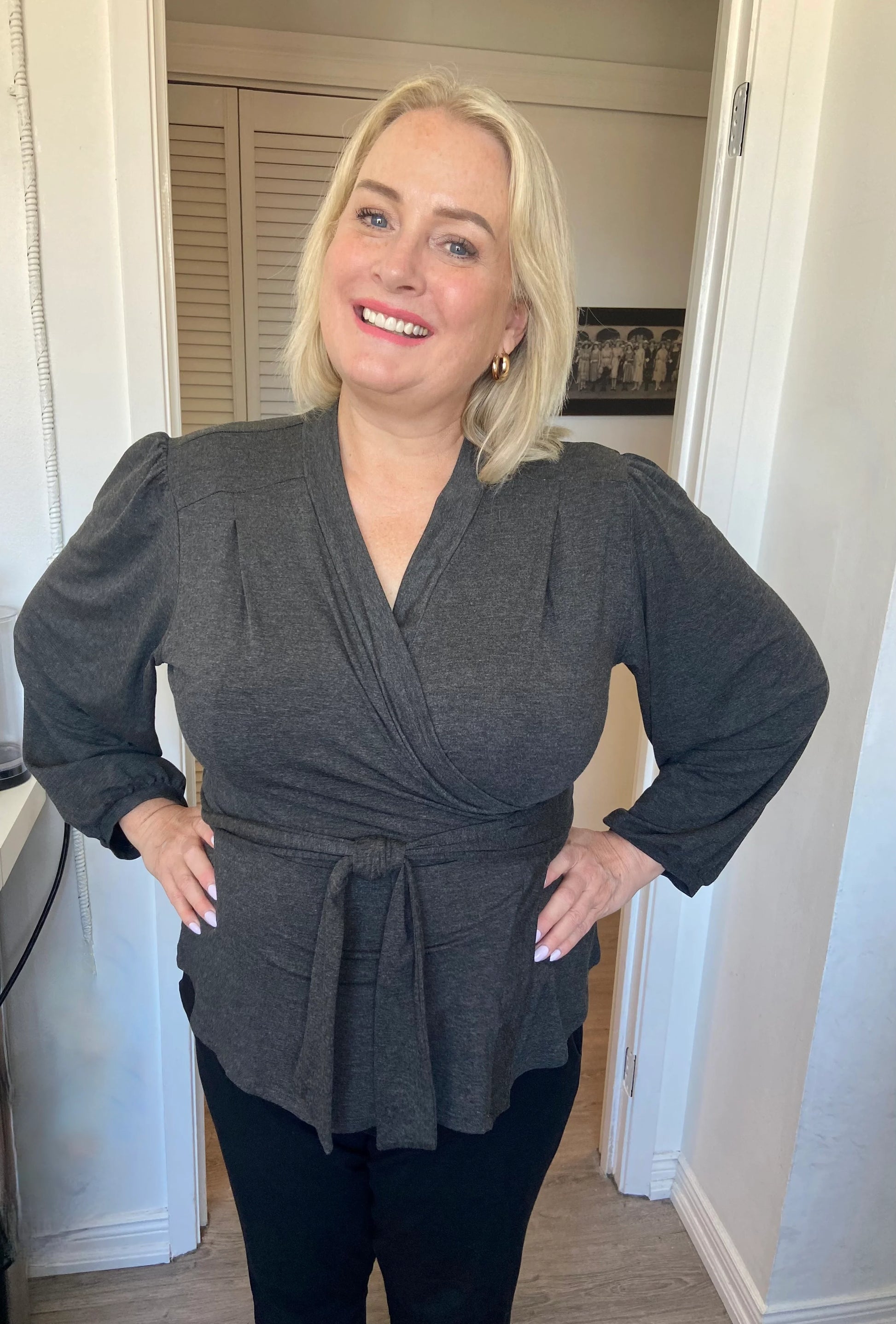 A woman wearing the Heather Black Bamboo Wrap Top from Dotty, with bracelet length sleeves and adjustable V-neckline. She is wearing it with a black pants and standing in a doorway. 